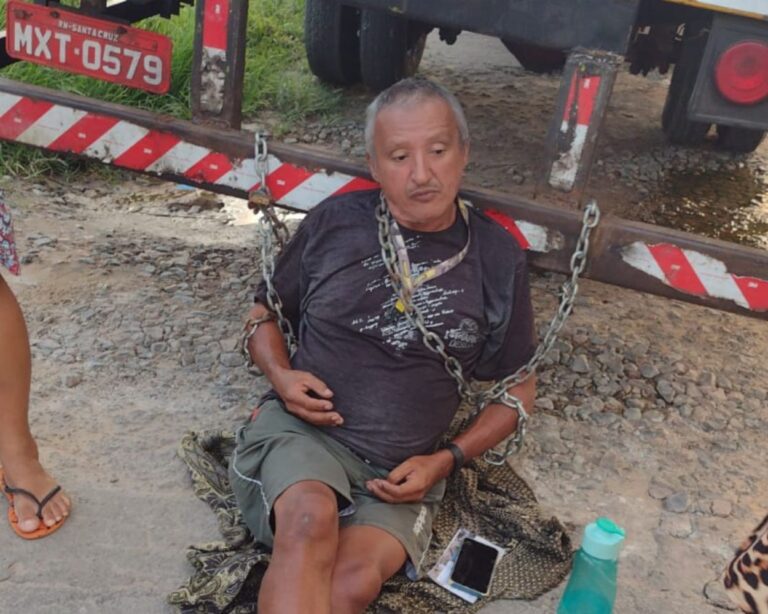 Imagem do homem sentado, acorrentado na traseira do caminhão baú.