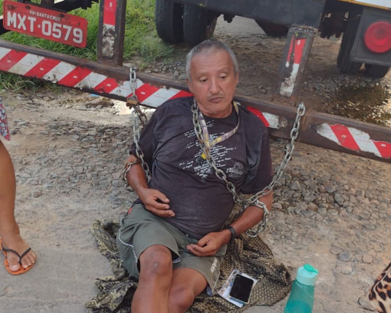 Imagem do homem sentado, acorrentado na traseira do caminhão baú.