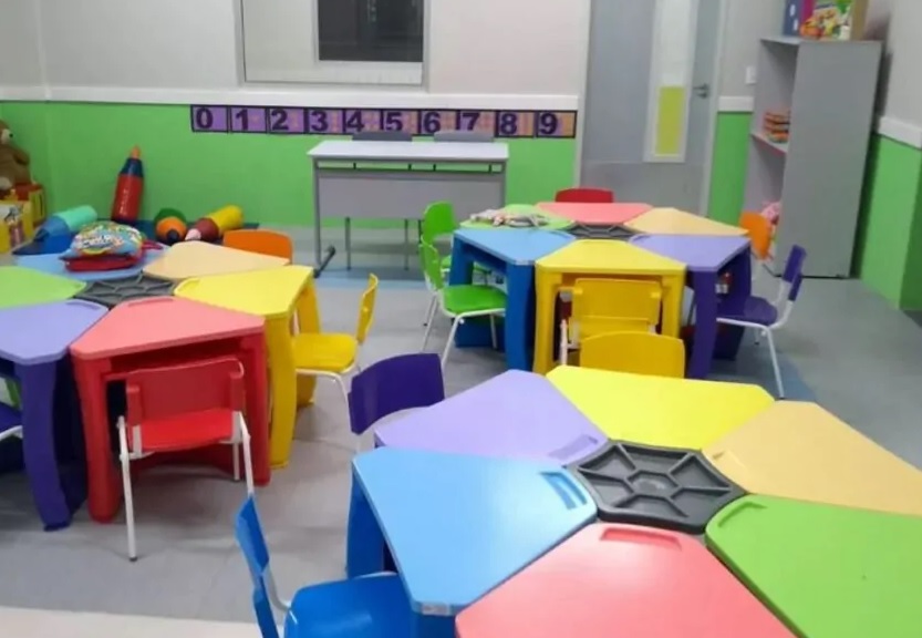 Imagem de sala de aula de creche com mesas e cadeiras coloridas.